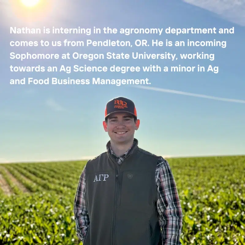 A man wearing a baseball cap and vest standing in front of a field with green crops. Transcribed Text: Nathan is interning in the agronomy department and comes to us from Pendleton, OR. He is an incoming Sophomore at Oregon State University, working towards an Ag Science degree with a minor in Ag and Food Business Management.