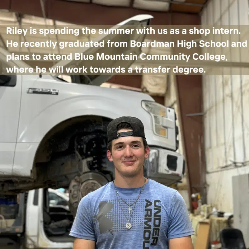 A young man standing in front of an elevated truck in a garage setting. Transcribed Text: Riley is spending the summer with us as a shop intern. He recently graduated from Boardman High School and plans to attend Blue Mountain Community College, where he will work towards a transfer degree.